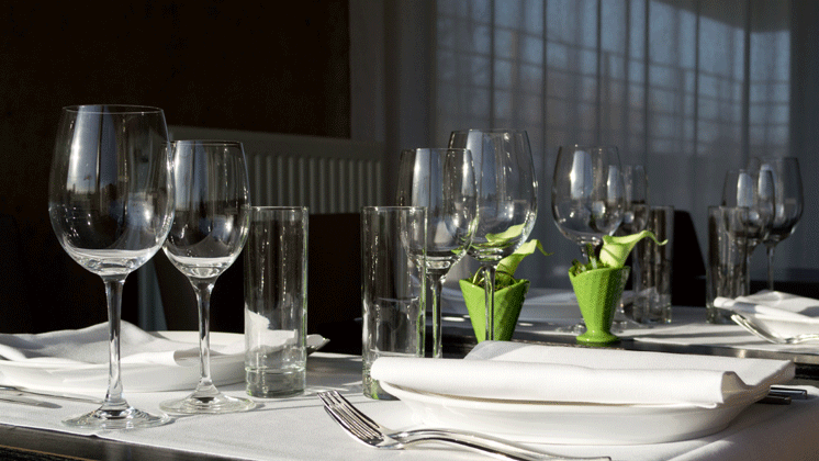 Photo d'une table de rstaurant dressée juste avant le repas. couverts en argent, assiètes et serviettes blanches, un verrre à eau et deux verres a vin.